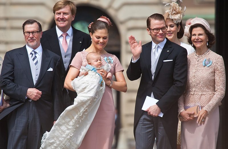 Royal family gathering at a christening ceremony with women wearing elegant pastel hats and fascinators, mother holding baby in white christening gown