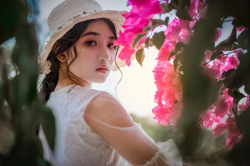 Woman wearing a cream straw hat with lace dress surrounded by pink bougainvillea flowers, elegant formal occasion styling