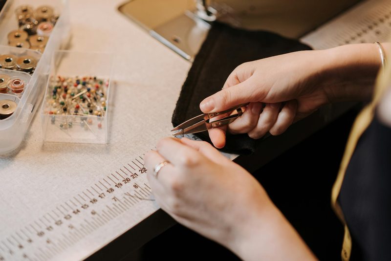 Close-up of a seamstress using sewing nippers to trim fabric, surrounded by pins, bobbins, and measuring tools in a millinery workshop