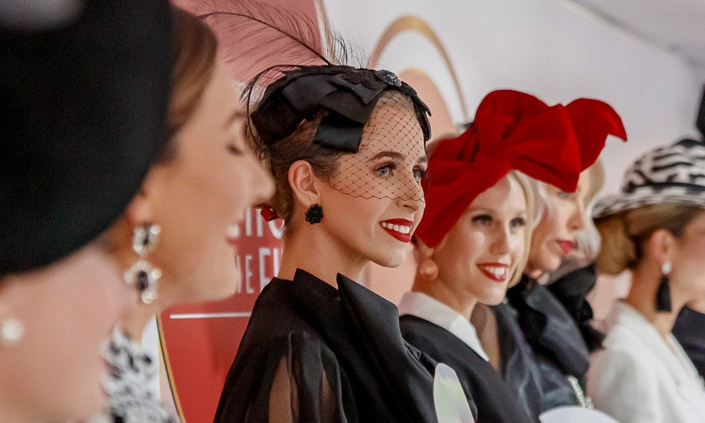 Spring racing millinery close-up featuring black veil fascinator with feathers and bold red bow headpiece